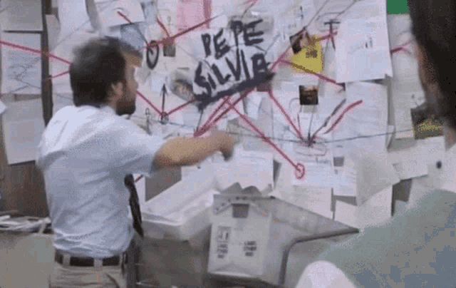 A scene from a television show featuring a man pointing at a chaotic board covered in papers, with red strings connecting different documents, emphasizing a frenzied investigation.